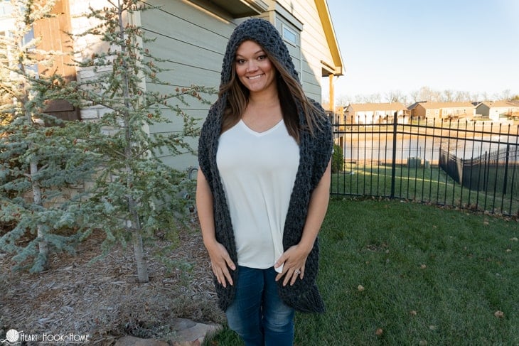 Smiling female model wearing a white t-shirt and jeans and a dark gray example of the Scrumptious Scoodie Hooded Scarf Crochet Pattern in a front yard setting.