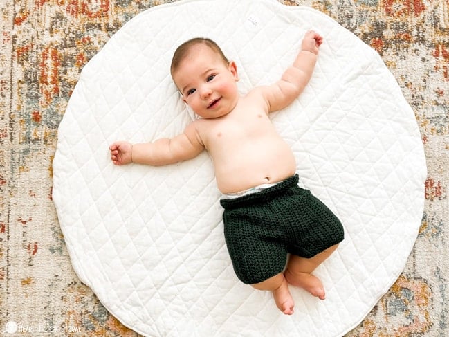 Infant model wearing an example of the Child Size Crochet Shorts in the “Ivy” colorway of Comfy Worsted yarn. The baby is laying on its back on a white circular rug. 