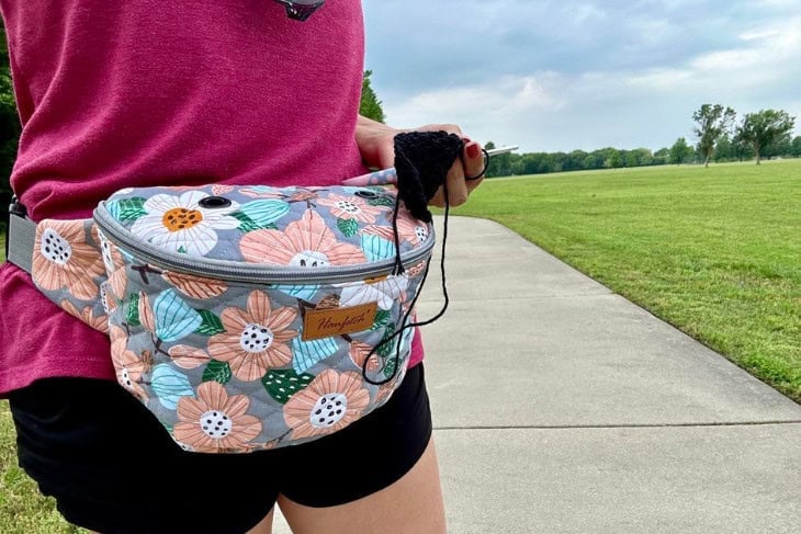 Flower and Gray yarn fanny pack on a female model holding a crochet hook and a sample of a crochet swatch in black yarn.